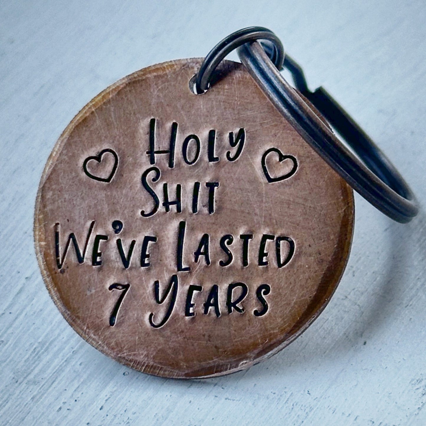 A bronze-colored copper keychain with the text 'HOLY SHIT WE'VE LASTED 7 YEARS' stamped on it, along with two heart shapes.