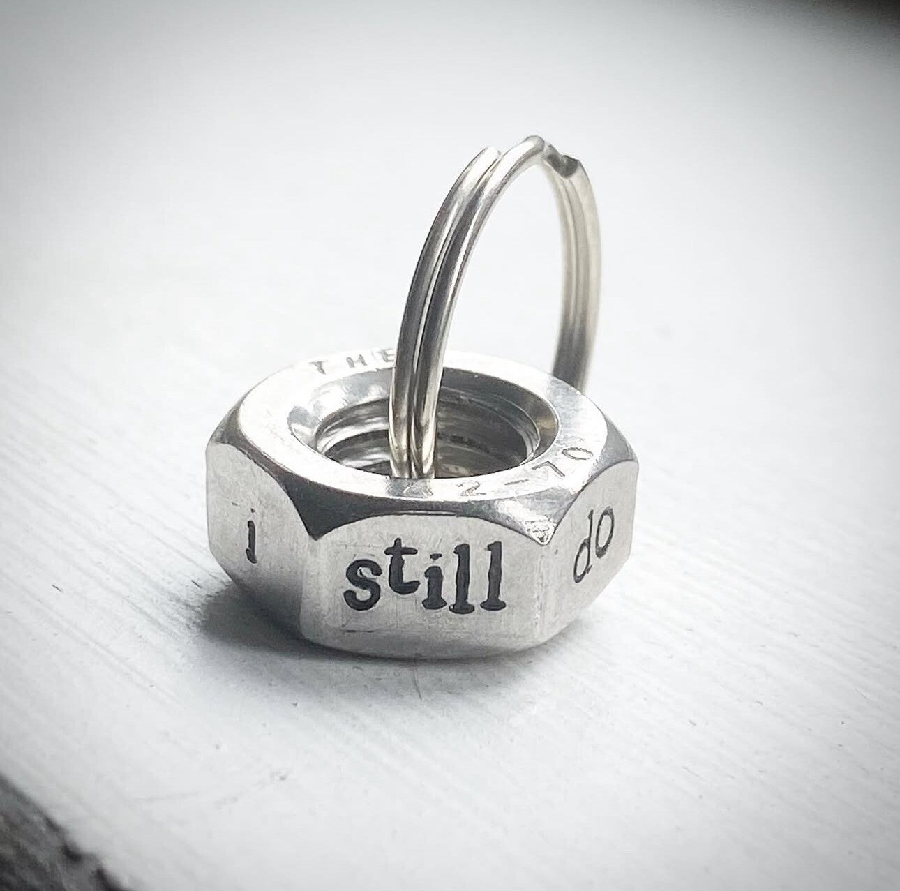A silver-colored hex nut keyring with the phrase 'I still do' stamped on it.