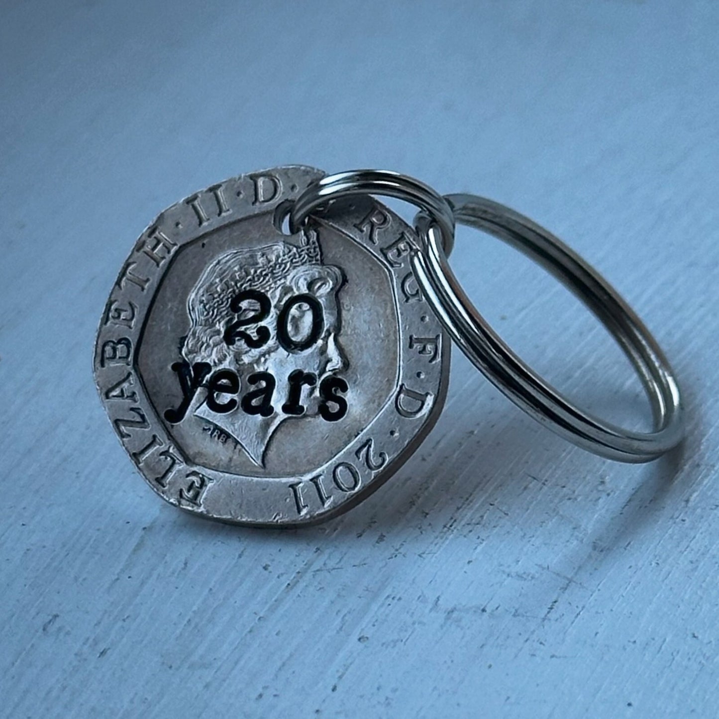 A silver-colored keychain featuring a hand-stamped 20p coin with '20 years' and additional text on it.