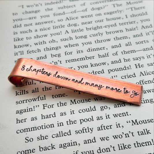 A bronze-colored metal bookmark with the inscription '8 CHAPTERS DOWN and MANY MORE TO GO' stamped on it