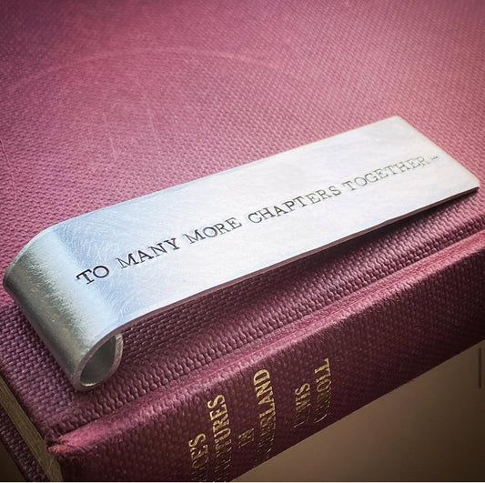 A silver aluminum bookmark with the inscription 'TO MANY MORE CHAPTERS TOGETHER' stamped on it.