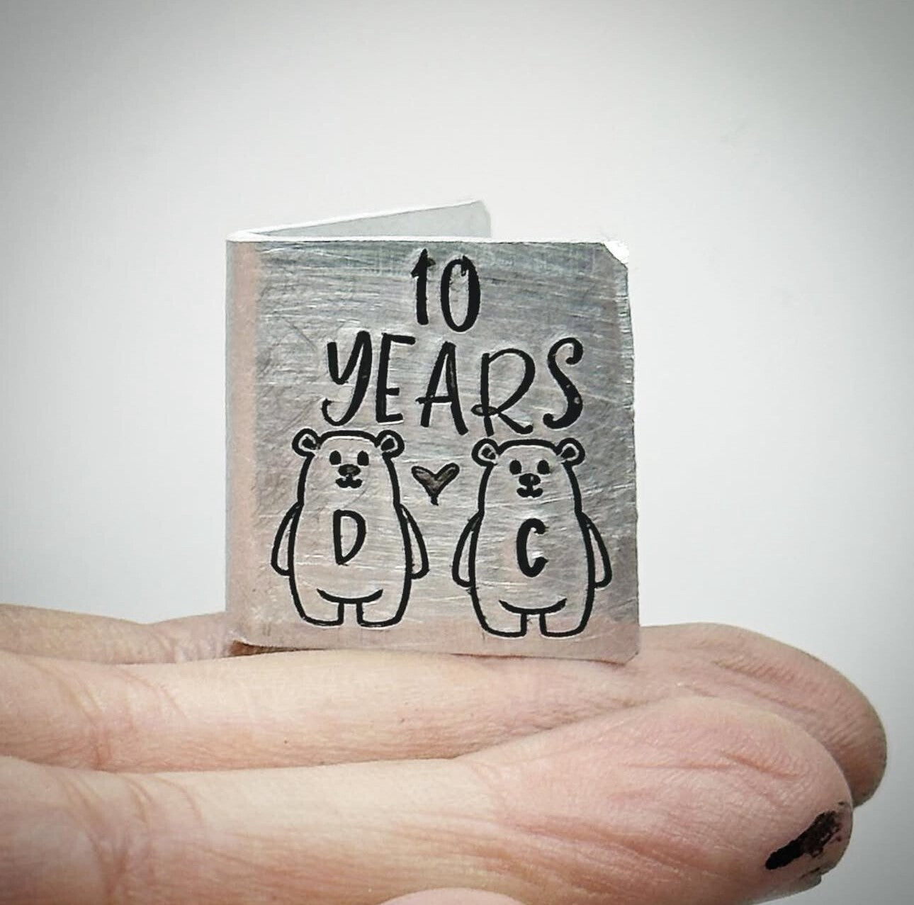 A hand stamped aluminium wedding anniversary card with '10 YEARS' and initials 'D C' stamped on the front, alongside a heart and two bears.