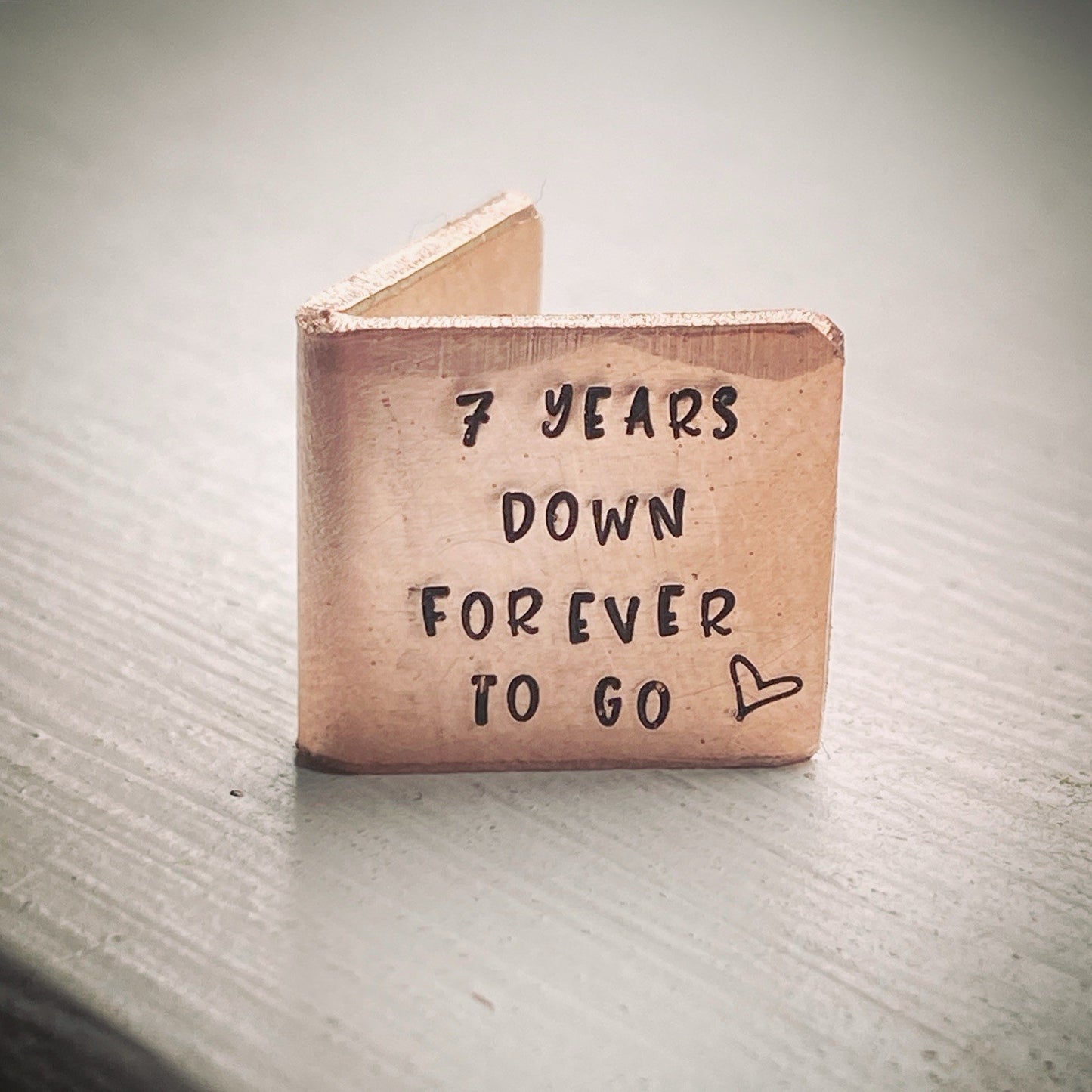 a small copper-colored metal card with the inscription '7 YEARS DOWN FOREVER TO GO' and a heart symbol.