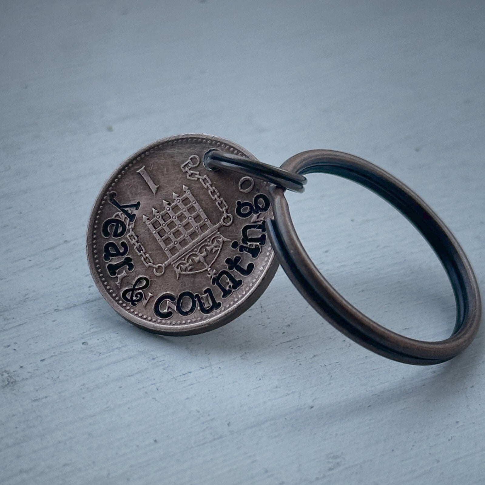 A keychain featuring a hand-stamped 1p coin with '1 year and counting' inscribed on it.