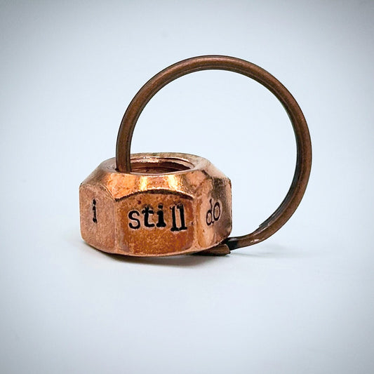 A copper hex nut keyring with the phrase 'I still do' stamped on it.