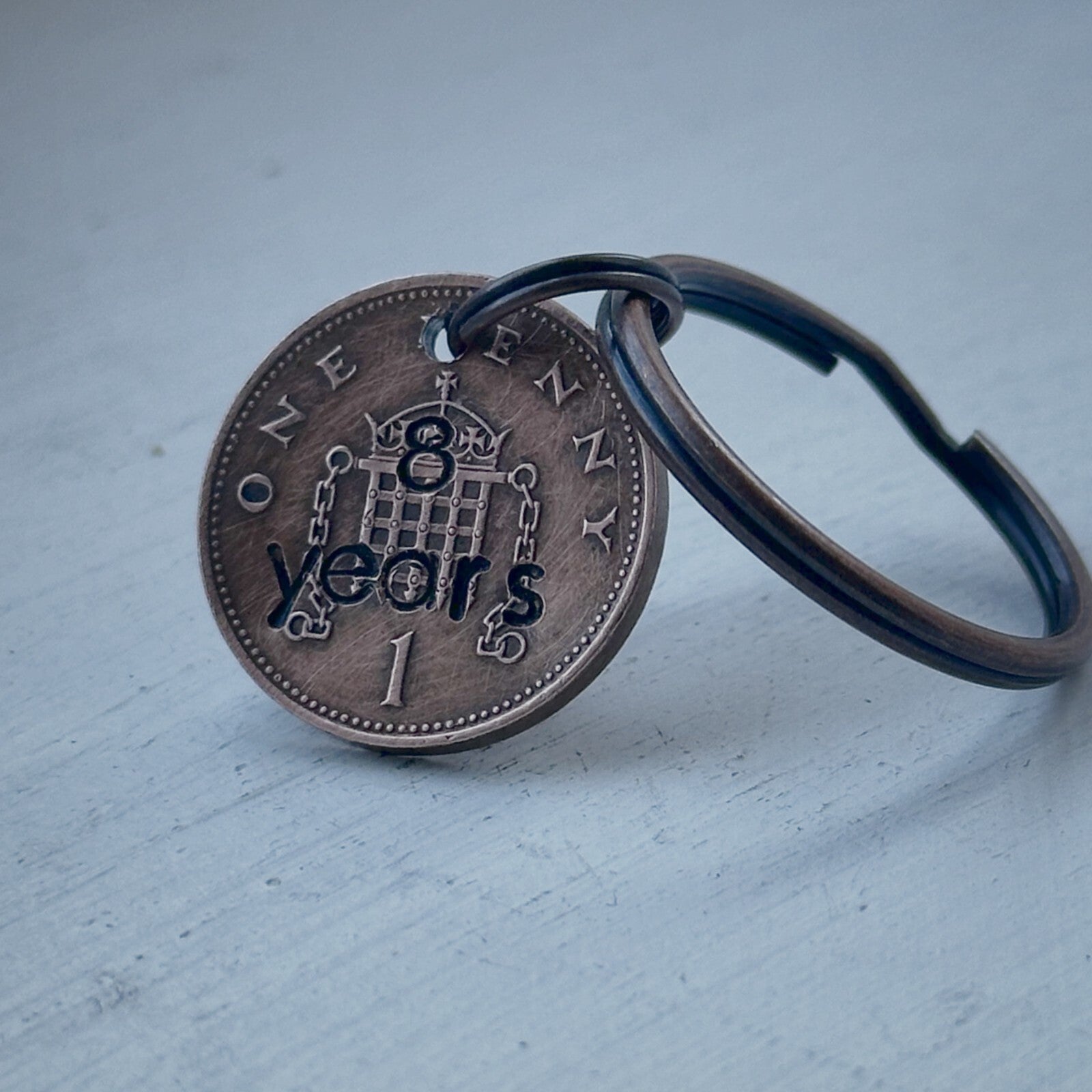 A bronze-colored keychain featuring a hand-stamped 1p coin with '8 years' and intricate designs, attached to a keyring.