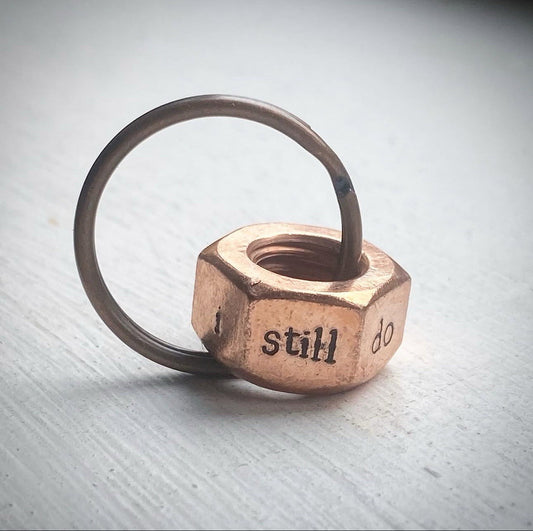 A copper hex nut keyring with 'I STILL DO' stamped on the sides.