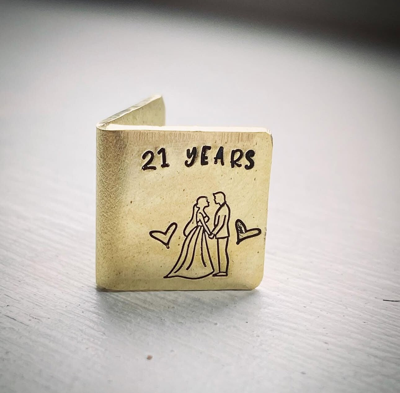 A personalized miniature brass card with '21 YEARS' and an illustration of a couple embracing, along with heart shapes.